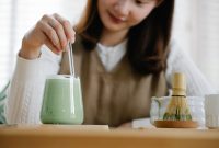 A woman stirs an iced matcha latte with a straw, using a traditional matcha whisk.