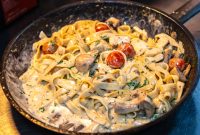 aap-creamy-tuscan-chicken-pasta-recipe-1790871343 Delicious creamy fettuccine pasta with mushrooms and tomatoes in a skillet, perfect for Italian cuisine lovers.