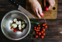 aap-cucumber-salad-recipes-for-hot-summer-days-1791295186 Hands preparing fresh vegetable salad with cherry tomatoes, cucumbers, and radishes on a wooden board.