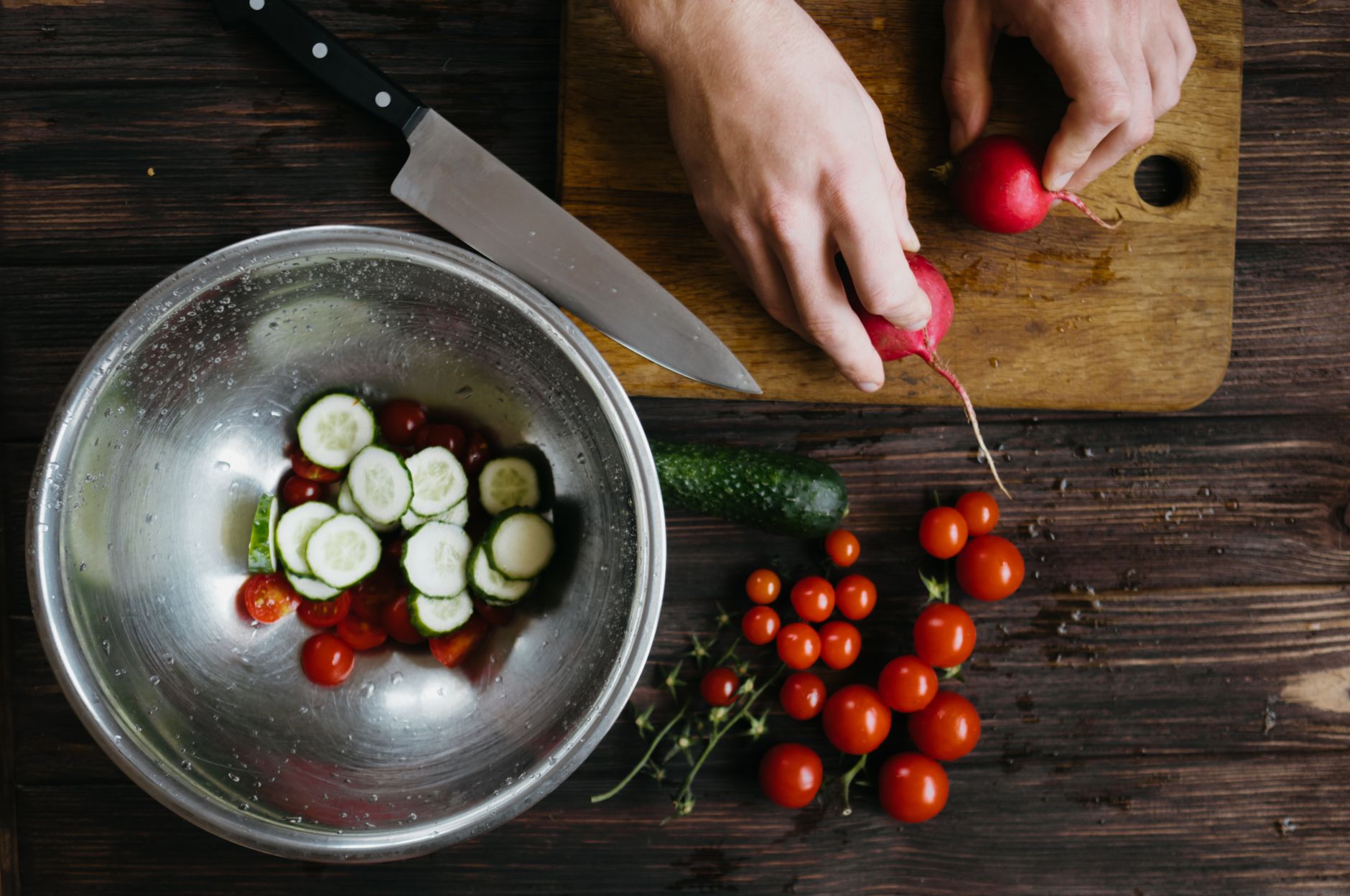 Hands preparing fresh vegetable salad with cherry tomatoes, cucumbers, and radishes on a wooden board.