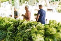 Vibrant display of green vegetables at an outdoor market with blurred shoppers in the background.
