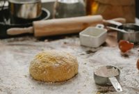 Ball of raw dough placed on table sprinkled with flour near rolling pin dishware and measuring cup in kitchen on blurred background
