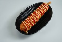 aap-korean-corn-dog-recipe-with-mozzarella-cheese-1791122297 Close-up of a Korean corn dog drizzled with ketchup and mustard on a black plate.