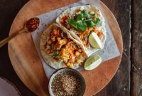 aap-traditional-mexican-street-food-recipes-to-make-at-home-1790864705 Top view of delicious Mexican tacos with fresh ingredients, lime, and salsa on a wooden plate.